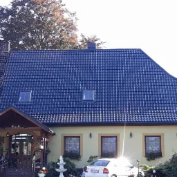 Dom z nowym, ciemnogranatowym dachem z dachówki ceramicznej z trzema oknami dachowymi, widoczny fragment starego dachu po lewej stronie, żółta elewacja, numer domu 10, biały samochód na podjeździe,...