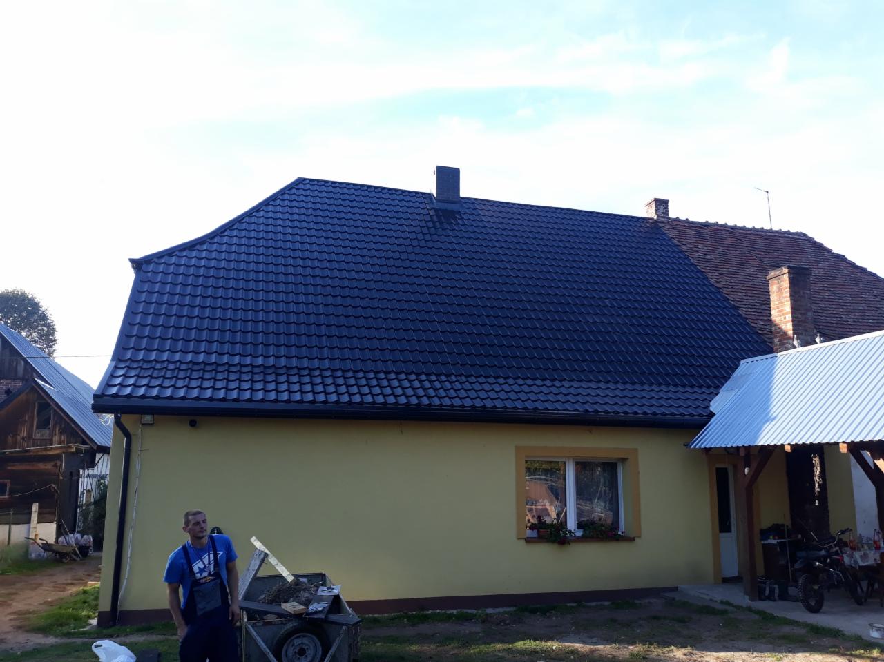 Dom jednorodzinny z nowym, ciemnym dachem z blachodachówki, widoczny pracownik w niebieskiej koszulce oraz przyczepka z narzędziami, w tle drugi budynek z ceglanym kominem.