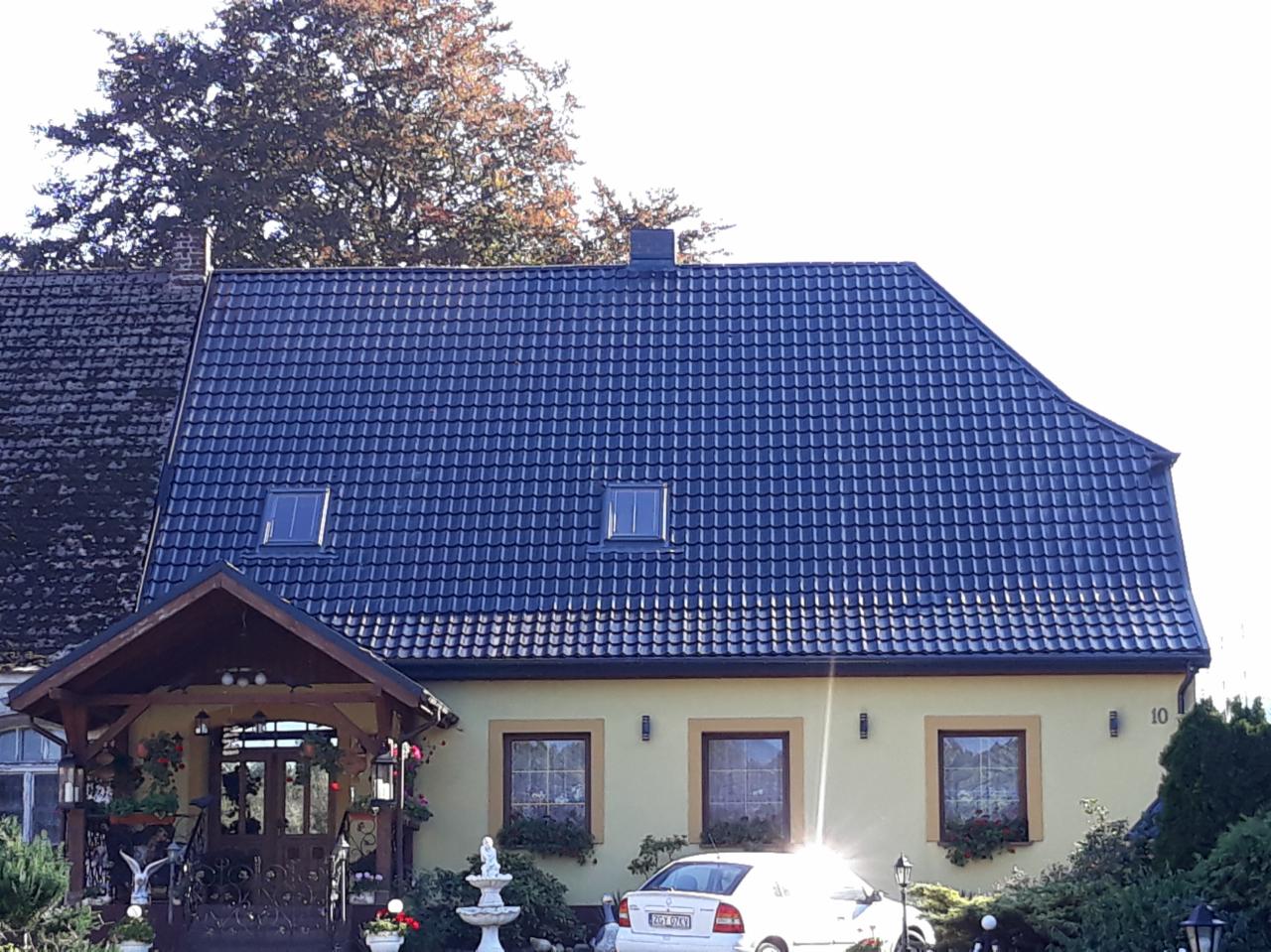 Dom z nowym, ciemnogranatowym dachem z dachówki ceramicznej z trzema oknami dachowymi, widoczny fragment starego dachu po lewej stronie, żółta elewacja, numer domu 10, biały samochód na podjeździe,...