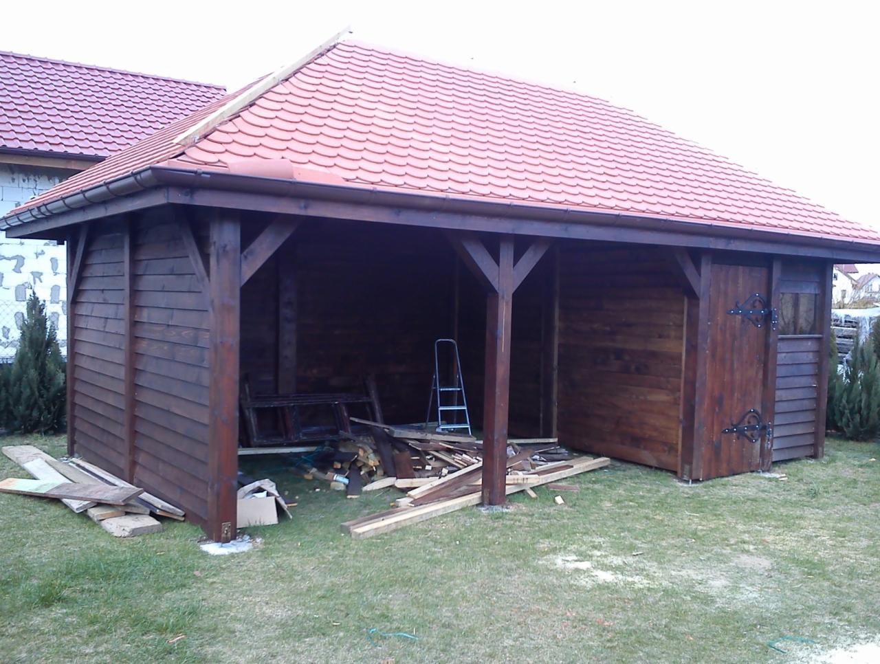 Drewniana altana z czerwonym dachem z dachówki ceramicznej w ogrodzie, z widocznymi elementami konstrukcyjnymi i resztkami materiałów budowlanych.