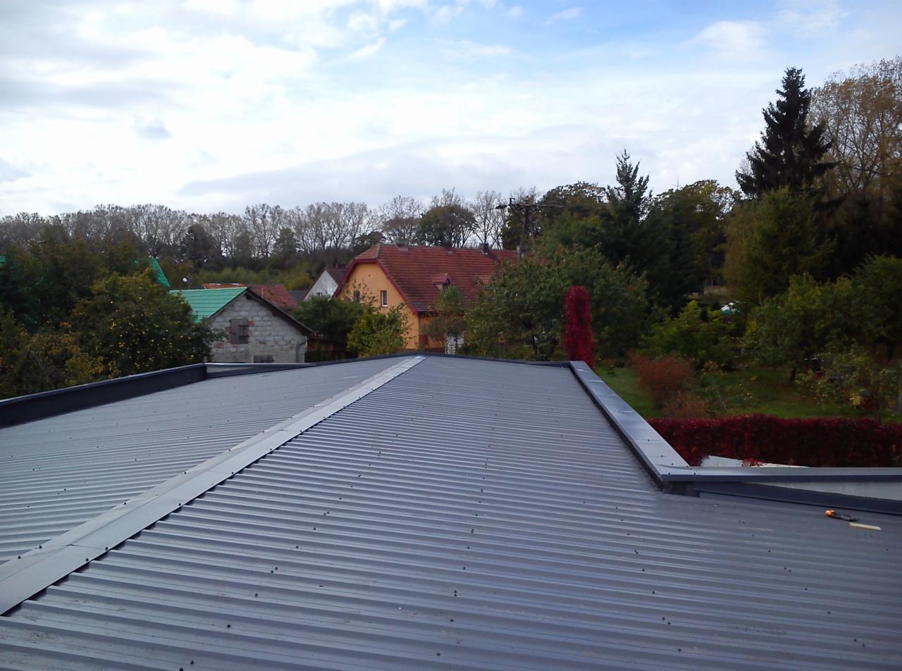 Nowy, szary, blaszany dach trapezowy z widocznymi łączeniami i obróbkami blacharskimi, na tle zabudowań mieszkalnych i zieleni.
