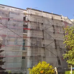 Budynek wielorodzinny w trakcie remontu elewacji, z rusztowaniami i siatkami ochronnymi, dwóch pracowników na rusztowaniu przy oknie.