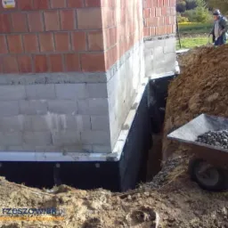Fundament budynku w trakcie izolacji przeciwwilgociowej z widoczną membraną kubełkową i drenażem z kamienia w wykopie, w tle osoba na zewnątrz.