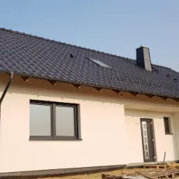 Nowoczesny dom z ciemną dachówką i oknami w kolorze antracytu w trakcie wykończenia, widoczne palety budowlane i fragment reklamy firmy.