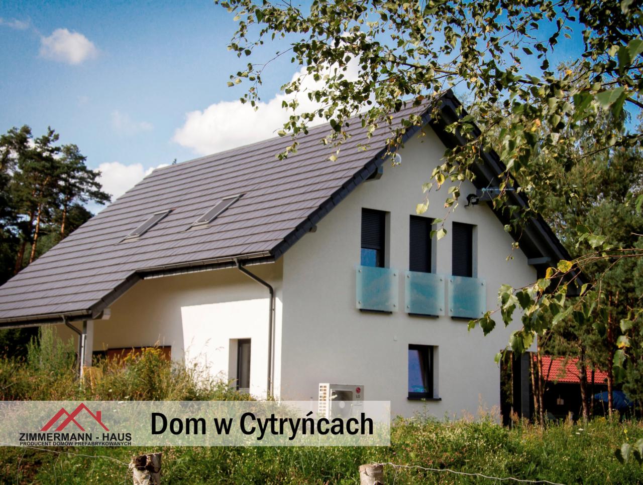 Gotowy dom jednorodzinny z szarą dachówką, jasną elewacją i balkonami z niebieskiego szkła, otoczony zielenią.