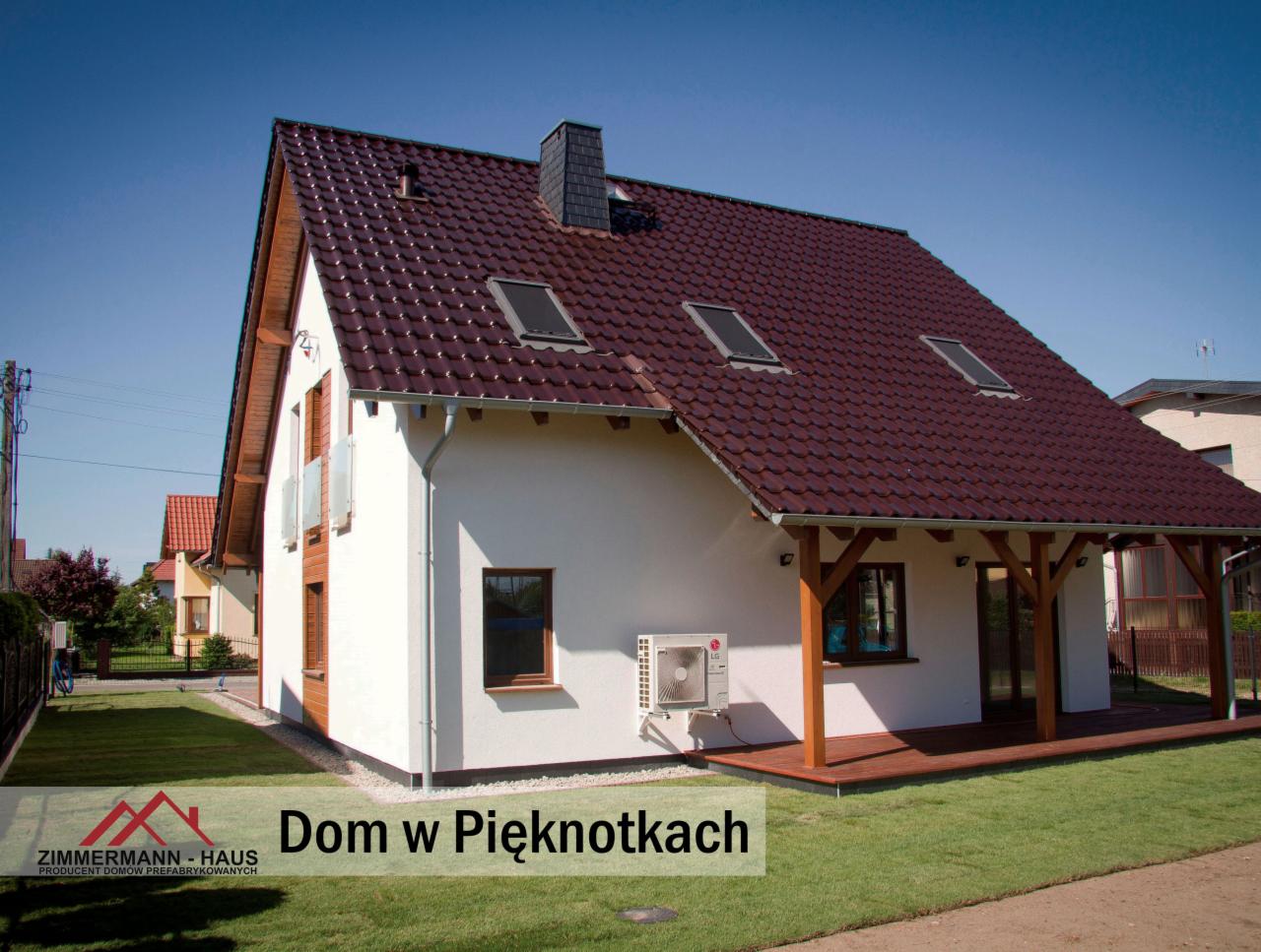 Gotowy dom z białymi ścianami, brązowym dachem z dachówką, drewnianą pergolą i klimatyzatorem na ścianie, widok z zewnątrz w słoneczny dzień.