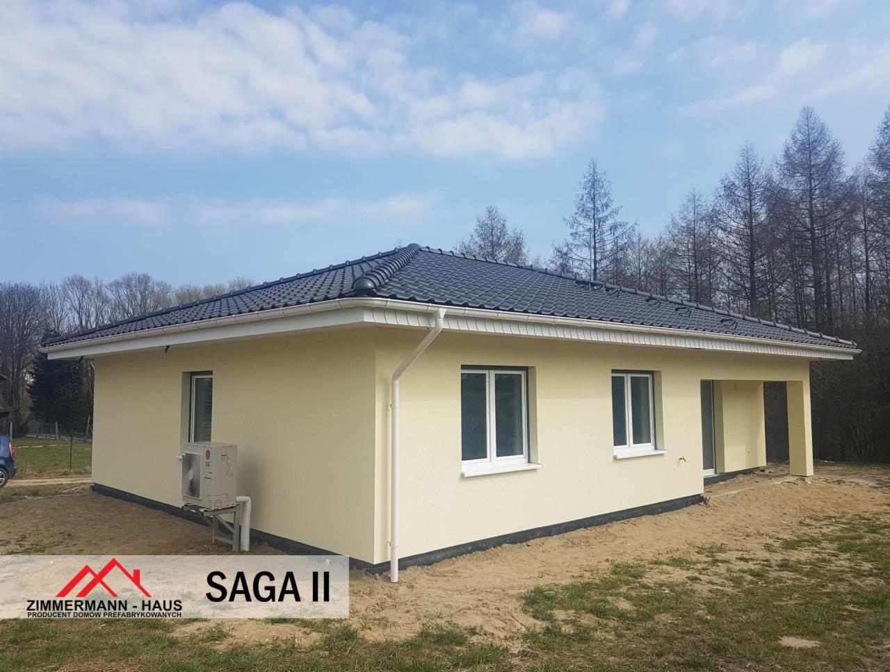 Gotowy parterowy dom z klimatyzacją zewnętrzną i ciemną dachówką, otynkowany na jasnożółto, widok z boku na tle drzew i błękitnego nieba.