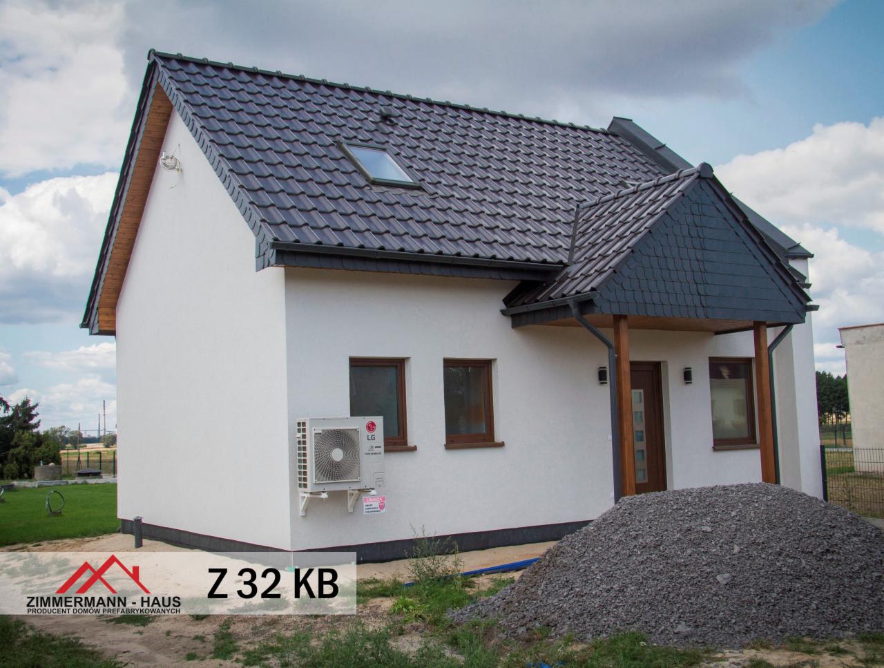 Gotowy, parterowy budynek z białym tynkiem, ciemną dachówką i drewnianymi elementami wykończeniowymi, z widoczną jednostką zewnętrzną klimatyzacji LG i stertą żwiru przed wejściem.