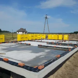 Wylany fundament pod dom murowany z izolacją pionową, obłożony bloczkami Ytong, widoczne wystające rury kanalizacyjne i cegły, w tle domy w budowie i słupy wysokiego napięcia.