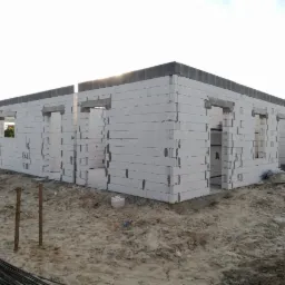 Wznoszenie ścian parterowego domu z betonu komórkowego Ytong, widoczne nadproża okienne i wieniec stropowy, teren budowy z nasypem piasku.