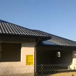 Nowy dom w budowie z czarną dachówką i rynnami, widok z zewnątrz przez ogrodzenie. Na ścianie budynku widoczna żółta tablica informacyjna.