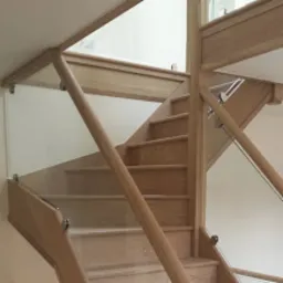 Jasne schody drewniane z balustradą szklaną, widok z dołu. Elementy drewniane o naturalnym odcieniu, gładkie, nowoczesny design. Minimalistyczne mocowania szkła.