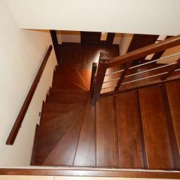 Jscarpentry - Kątowy widok z góry na ciemnobrązowe schody z drewna, z metalową poręczą. Widoczne stopnie skręcają, a w tle widać ciemną podłogę.