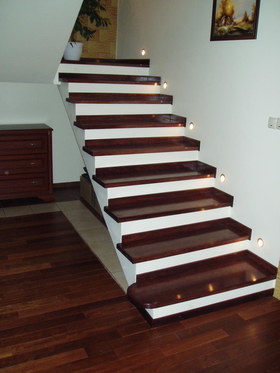 Schody z ciemnego drewna z białymi podstopnicami i oświetleniem punktowym w ścianie, widoczne drewniane podłogi i komoda w tle. Detal wykończenia wnętrza.