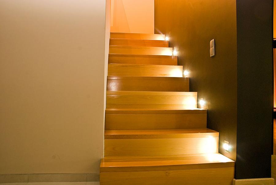 Jasne drewniane schody z dyskretnym oświetleniem LED wzdłuż stopni, na tle ściany w odcieniach beżu i brązu. Minimalistyczny design.