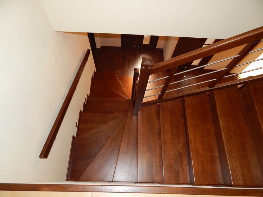 Kątowy widok z góry na ciemnobrązowe schody z drewna, z metalową poręczą. Widoczne stopnie skręcają, a w tle widać ciemną podłogę.
