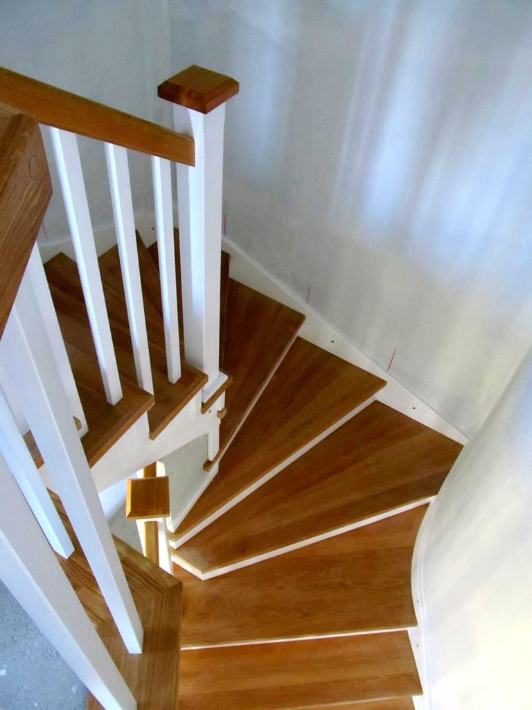 Kątowe ujęcie krętych schodów z drewna o ciepłym odcieniu, z białymi balustradami i drewnianym poręczem, w jasnym, minimalistycznym wnętrzu.