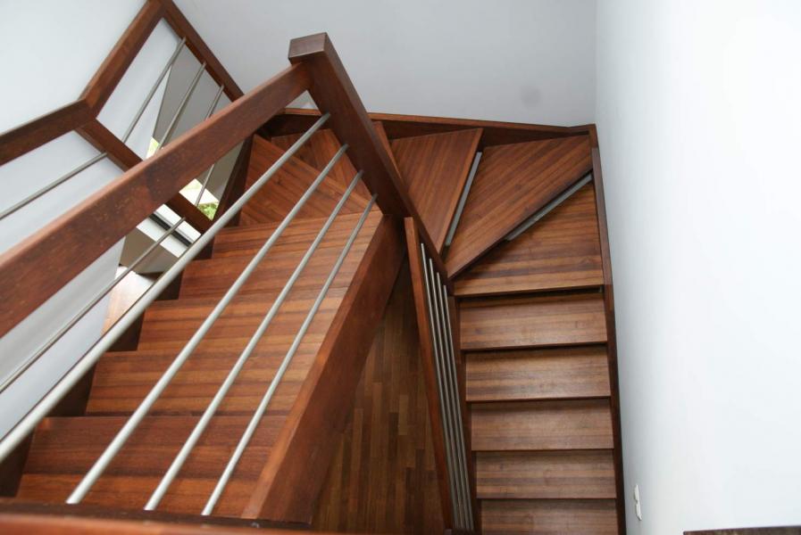 Drewniane schody z metalowymi poręczami w nowoczesnym domu. Ujęcie z dołu, eksponujące detale wykończenia i naturalny odcień drewna. Widoczne stopnie i fragment ściany.