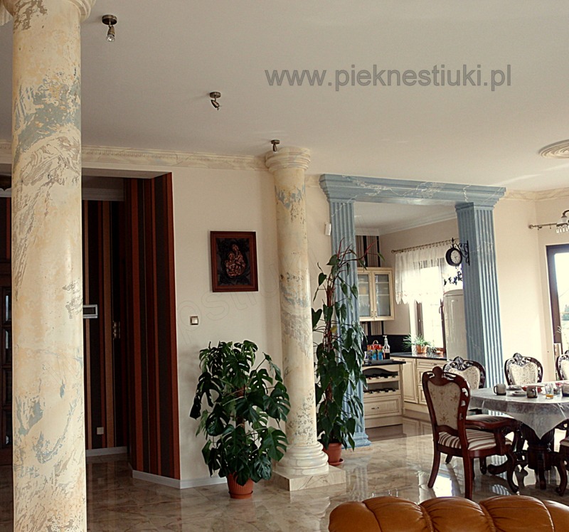 Salon z kolumnami wykończonymi stiukiem imitującym marmur, widoczne rośliny doniczkowe i błyszcząca podłoga.