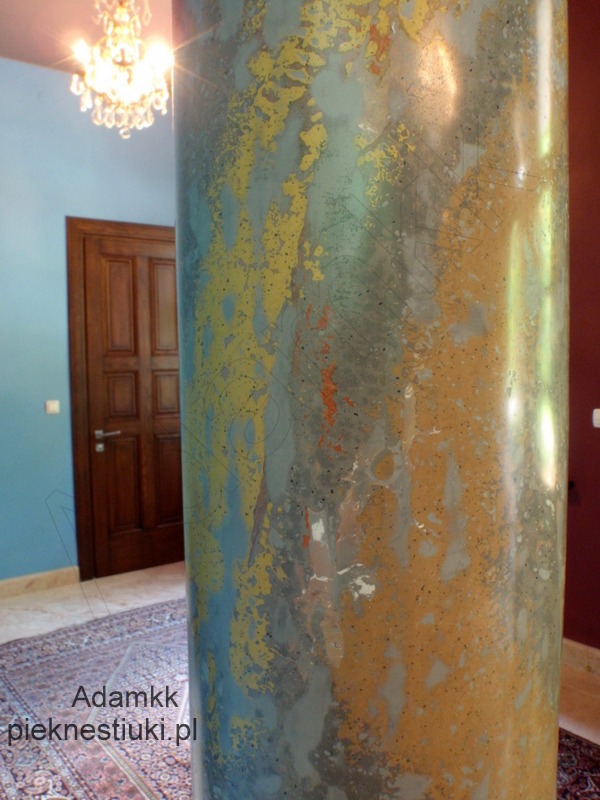 Kolumna wewnątrz pomieszczenia wykończona stiukiem imitującym marmur w odcieniach złota, błękitu i szarości, z widocznym wzorem marmoryzacji, na tle drzwi i kryształowego żyrandola.