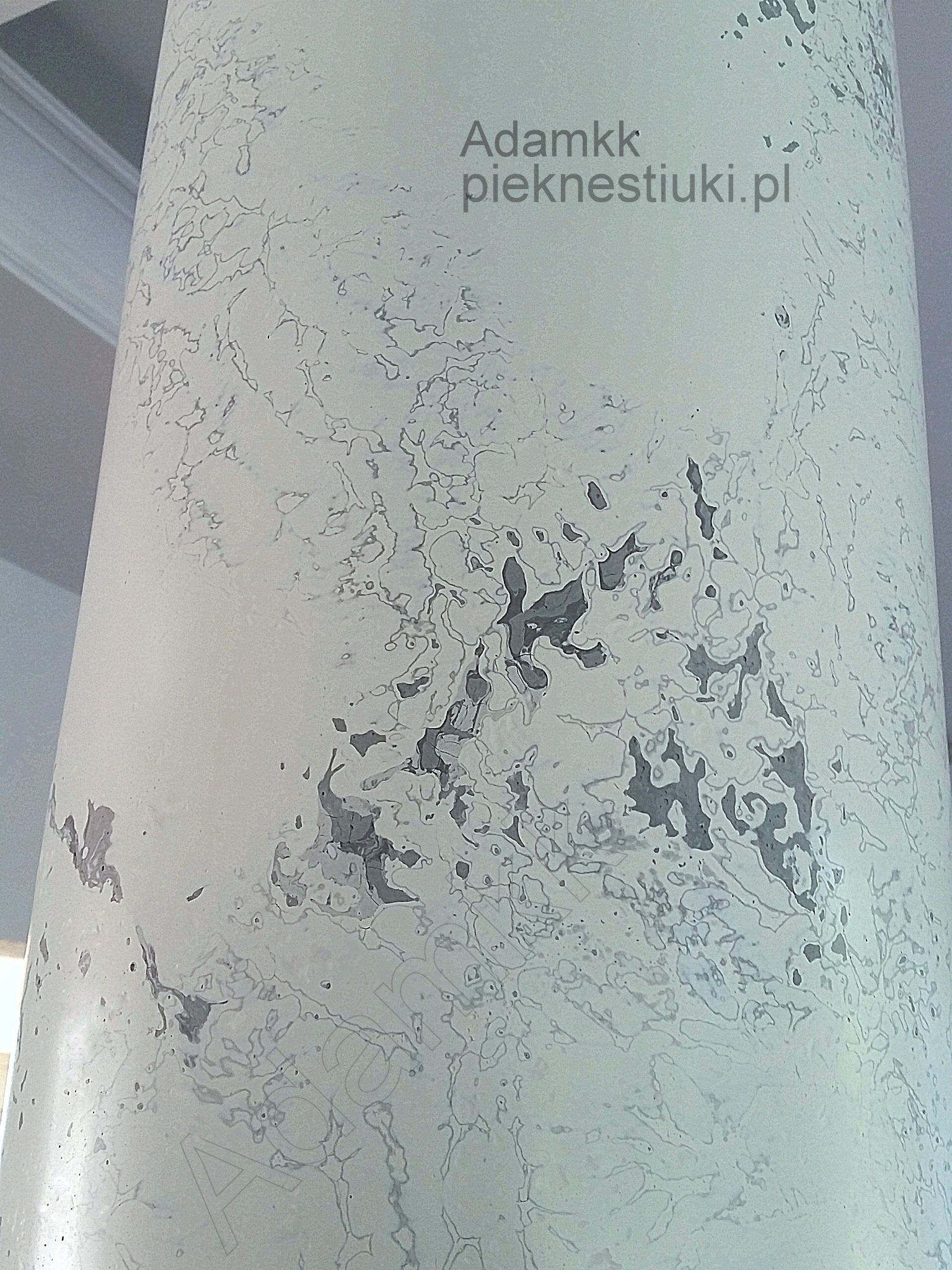 Kolumna wykończona stiukiem porcelanowym z efektem marmoryzacji, widoczne nieregularne wzory i szare plamy imitujące kamień, w górnej części widoczny adres strony internetowej.