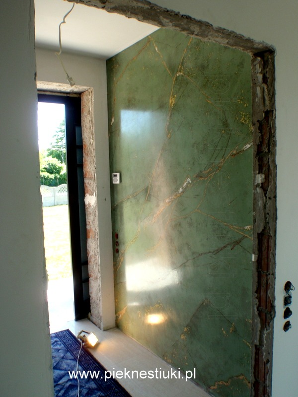 Ściana wykończona stiukiem w odcieniach zieleni imitującym marmur, z widocznymi złotymi żyłkami, w trakcie prac wykończeniowych; surowy fragment ściany obok drzwi.