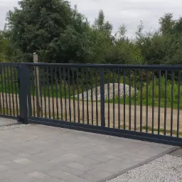 Przesuwna, metalowa brama wjazdowa w kolorze grafitowym z pionowymi prętami, widok z zewnątrz na tle zieleni i sterty kamieni.