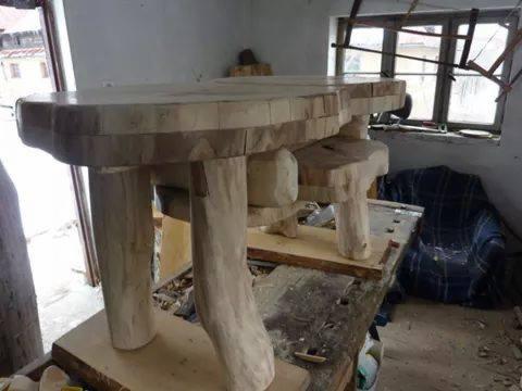 Surowy stół z drewna w warsztacie, z nogami z gałęzi i blatem z nieregularnych kawałków, w tle okno i narzędzia.