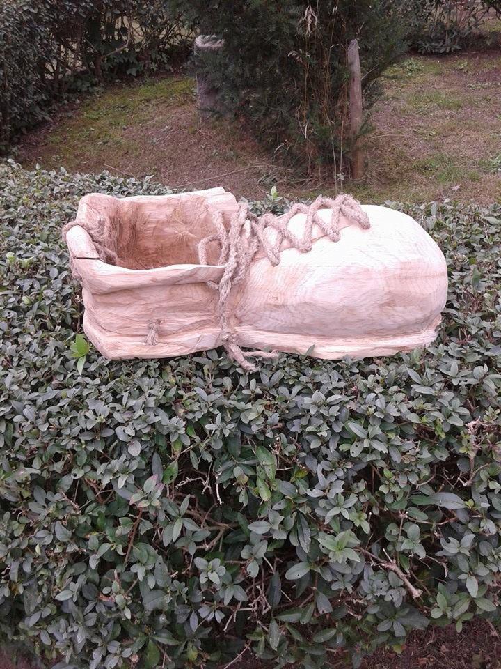 Ręcznie rzeźbiona, drewniana donica w kształcie buta, umieszczona na krzewie w ogrodzie. Sznurowadła wykonane z grubego sznura. Unikalny element dekoracyjny.