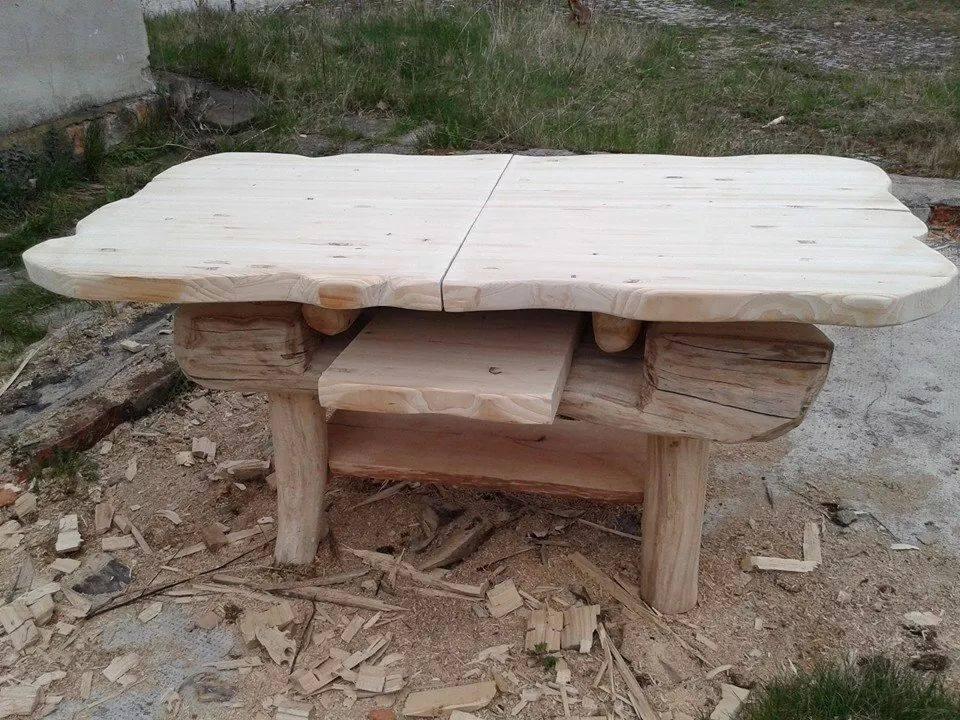 Surowy, drewniany stół z blatem z dwóch desek o nieregularnych krawędziach, wsparty na nogach z okrąglaków, otoczony wiórami.