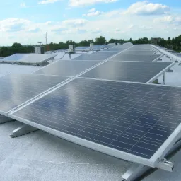 Rząd paneli fotowoltaicznych na płaskim dachu budynku przemysłowego, widoczne metalowe konstrukcje wsporcze i elementy wentylacji, w tle zieleń i błękitne niebo z chmurami.