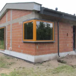 Surowy budynek z cegły z zamontowanymi oknami z drewnianymi ramami. Widoczne orynnowanie i fundamenty. Na zewnątrz budynku widać osoby.