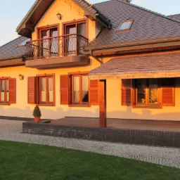 Elewacja domu jednorodzinnego z brązowymi oknami i okiennicami w promieniach zachodzącego słońca. Widoczny balkon z kutą balustradą i dach z dachówki.