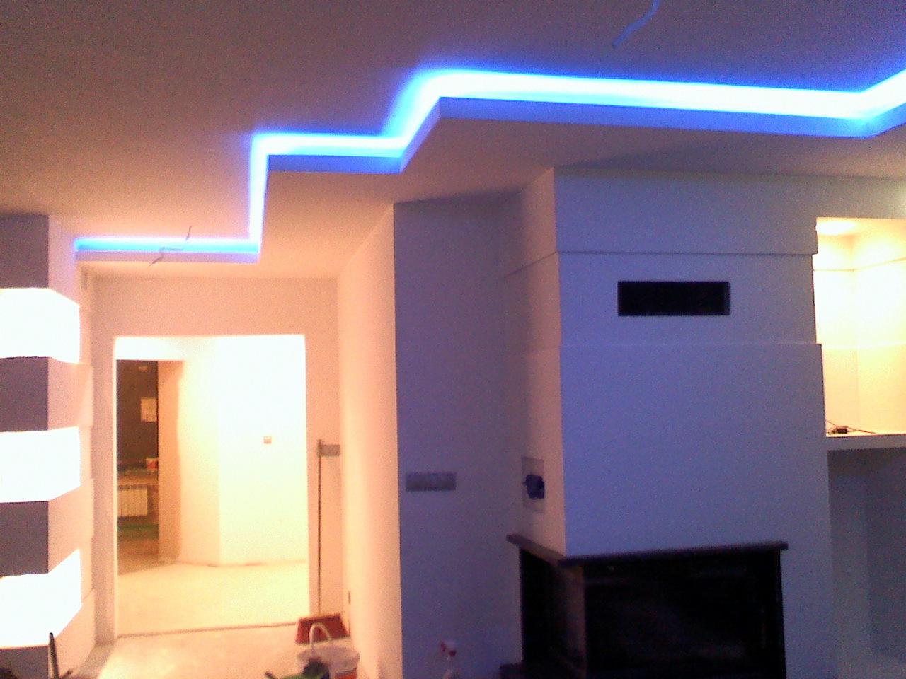 Nowoczesne wykończenie wnętrza z podwieszanym sufitem i liniowym oświetleniem LED w kolorze niebieskim. Widoczny fragment kominka i przejście do innego pomieszczenia.