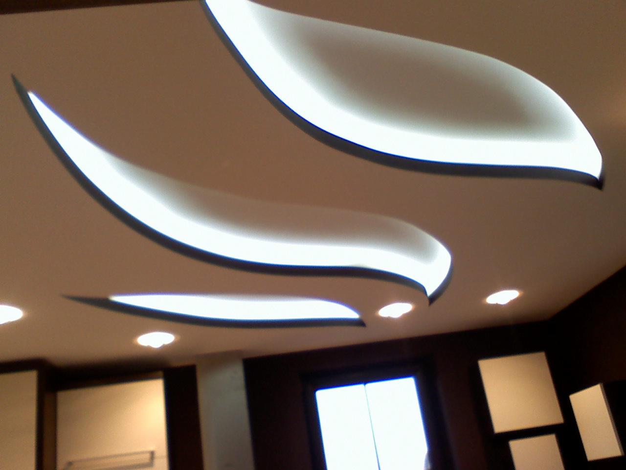 Nowoczesny sufit z dekoracyjnymi wnękami oświetleniowymi w kształcie liści i punktowym oświetleniem, widok na fragment pomieszczenia z oknem.