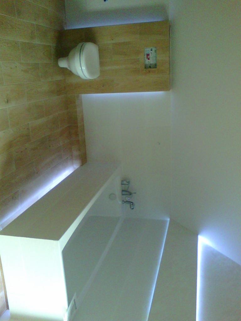 Nowoczesna łazienka z podświetleniem LED: drewnopodobne płytki, biała armatura i minimalistyczny design. Ujęcie z nietypowej perspektywy.