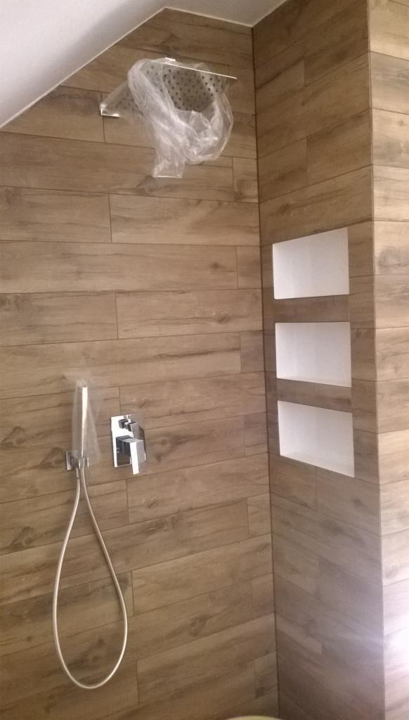 Wykończona kabina prysznicowa z drewnopodobnymi płytkami, chromowaną armaturą i wnękami w ścianie, zabezpieczona folią ochronną na czas remontu.