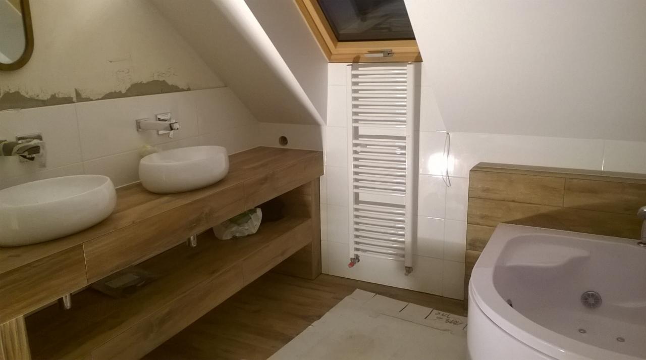 Łazienka na poddaszu z drewnianą szafką pod umywalki nablatowe, grzejnikiem drabinkowym pod oknem dachowym i obudową wanny z drewnopodobnych paneli, widoczne ślady po kleju na ścianie.