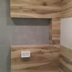 Ściana łazienki wyłożona płytkami imitującymi drewno, z widoczną wnęką i przygotowanymi otworami na armaturę oraz częściowo zamontowanym stelażem podtynkowym WC.