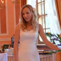 Panna młoda w koronkowej sukni ślubnej, z kwiatem we włosach, stojąca w elegancko udekorowanym wnętrzu sali weselnej, z widocznym stołem zastawionym ciastami i napojami.