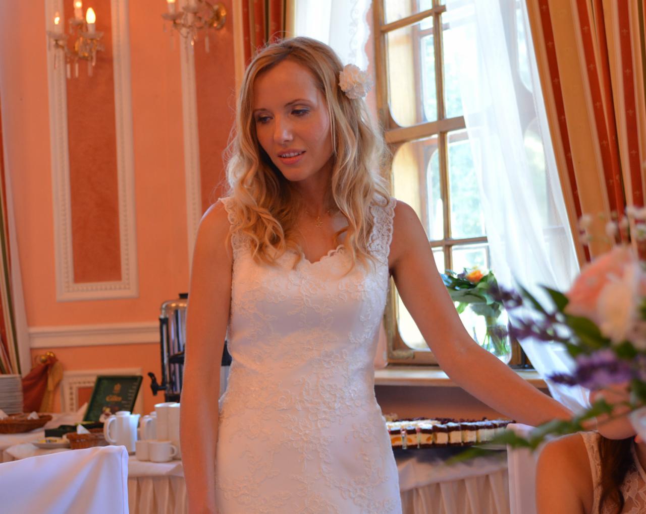 Panna młoda w koronkowej sukni ślubnej, z kwiatem we włosach, stojąca w elegancko udekorowanym wnętrzu sali weselnej, z widocznym stołem zastawionym ciastami i napojami.