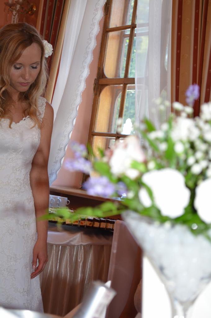 Panna młoda w koronkowej sukni spogląda w dół, obok okna z białymi firankami i stołu z przekąskami, bukiet kwiatów w szklanym wazonie na pierwszym planie.