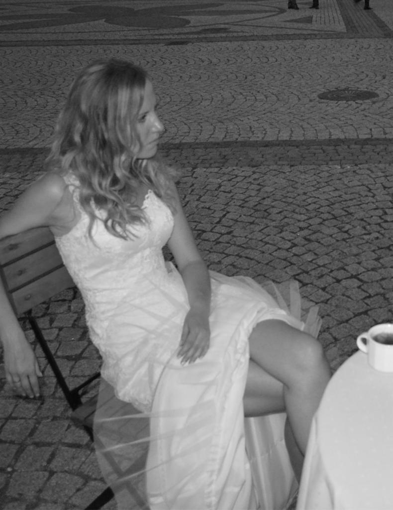 Czarno-biała fotografia panny młodej w sukni ślubnej siedzącej na krześle na zewnątrz, z filiżanką na stole obok.