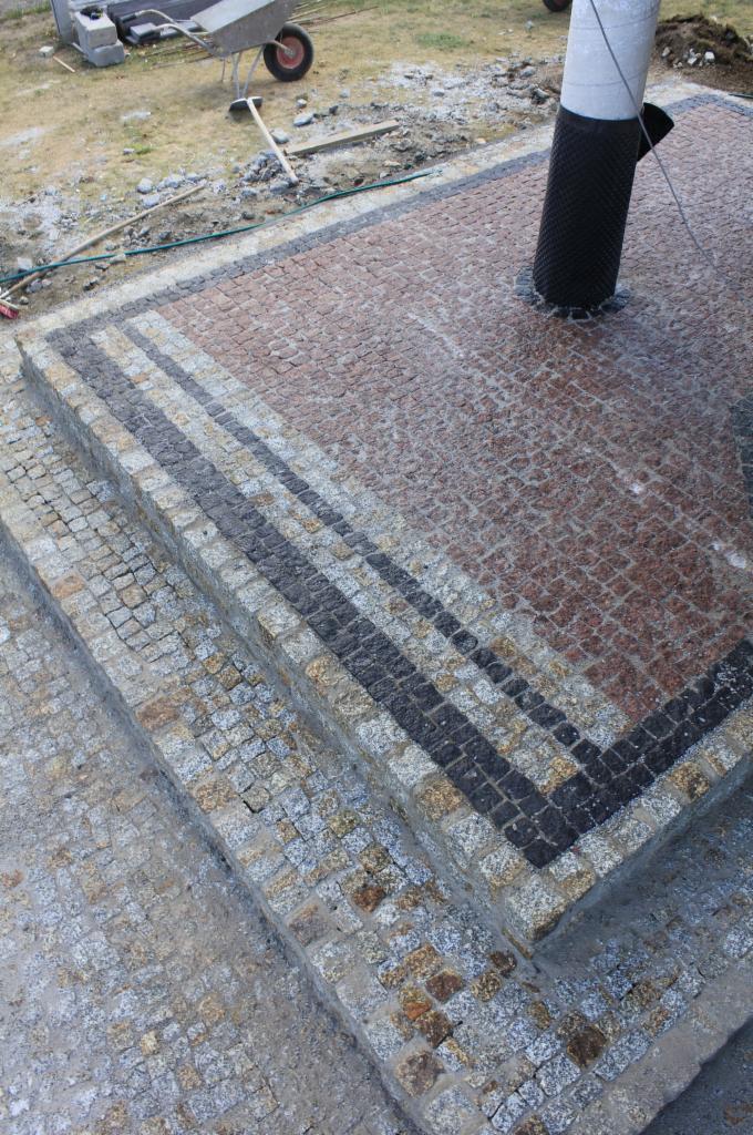 Schody i fragment nawierzchni z kostki granitowej w odcieniach szarości i czerwieni, z widocznym słupem i narzędziami brukarskimi w tle.