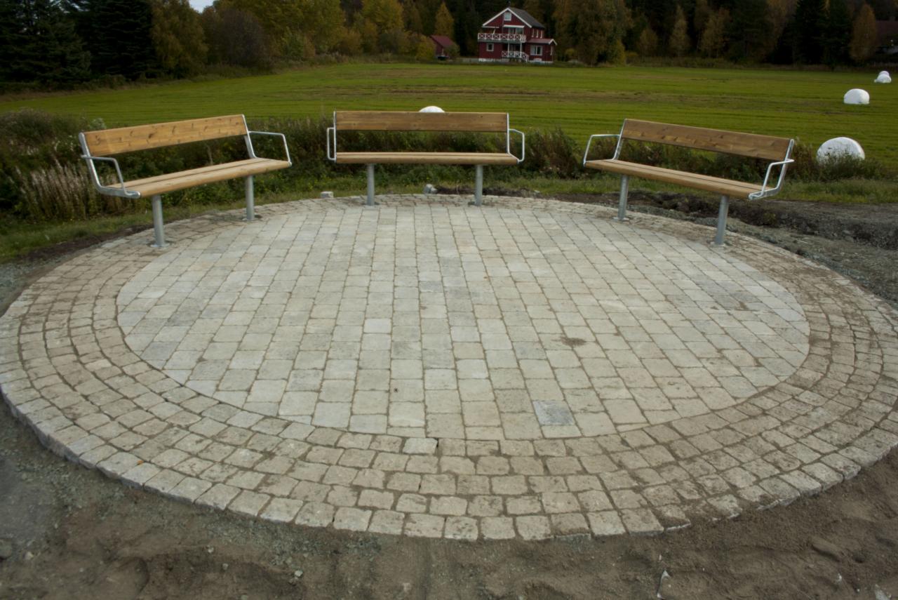Okazały, okrągły plac z jasnej kostki brukowej z trzema ławkami z drewnianym siedziskiem i metalowymi poręczami. W tle trawnik i czerwony dom.