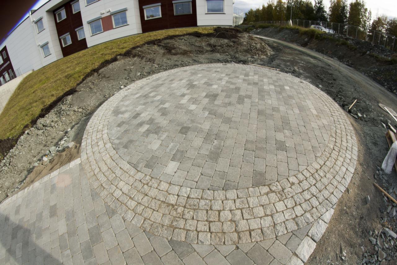 Okrągły plac z kostki granitowej w odcieniach szarości, otoczony pasem z jaśniejszej kostki, na tle budynku i wzniesienia. Widoczne narzędzia i materiały budowlane.