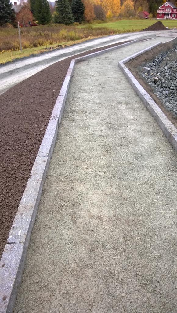 Ścieżka z podbudową i obrzeżami z kostki, przygotowana do ułożenia nawierzchni. Widok z perspektywy pieszego, w tle krajobraz jesienny i dom.