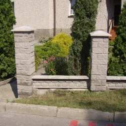 Niski murek z betonowych element&oacute;w imitujących cegłę, z filarami po bokach, oddzielający ogr&oacute;d od chodnika. W tle zieleń ogrodu i fragment budynku.