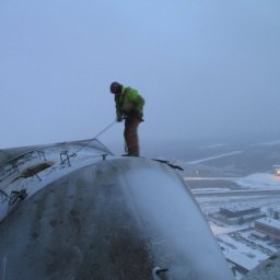ZAKŁAD USŁUG ALPINISTYCZNYCH RAFAŁ SENDELA - Pracownik w odblaskowej kurtce zabezpieczony linami, na szczycie metalowej konstrukcji pokrytej śniegiem, prawdopodobnie podczas prac wysokościowych lub naprawy dachu w zimowych warunkach.