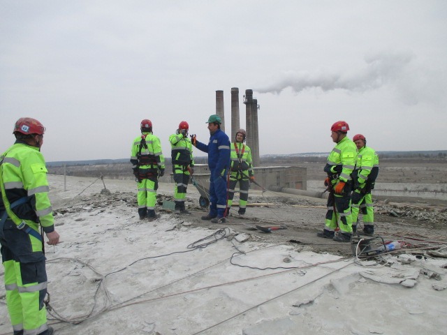Grupa pracowników w odblaskowych ubraniach i kaskach, zabezpieczonych linami, na zniszczonej powierzchni dachu, z kominami w tle i pochmurnym niebem.